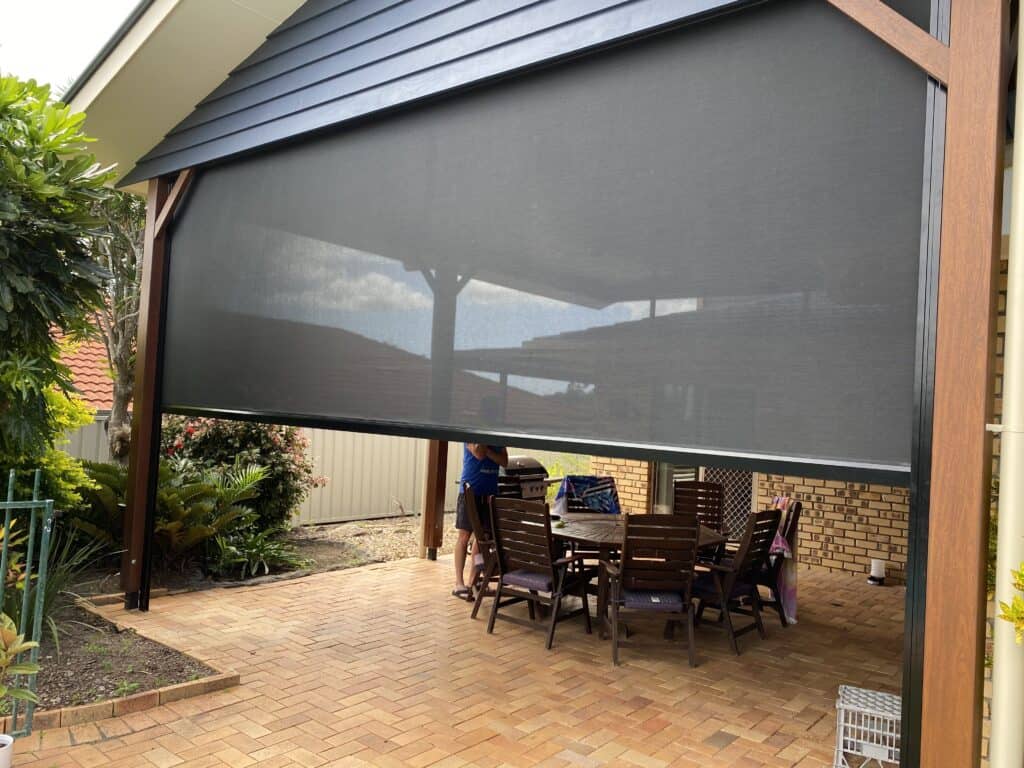 A covered outdoor patio area in Albany Creek features a wooden dining table and chairs, partially enclosed by a large black mesh screen. A person stands nearby, arranging items on the table.