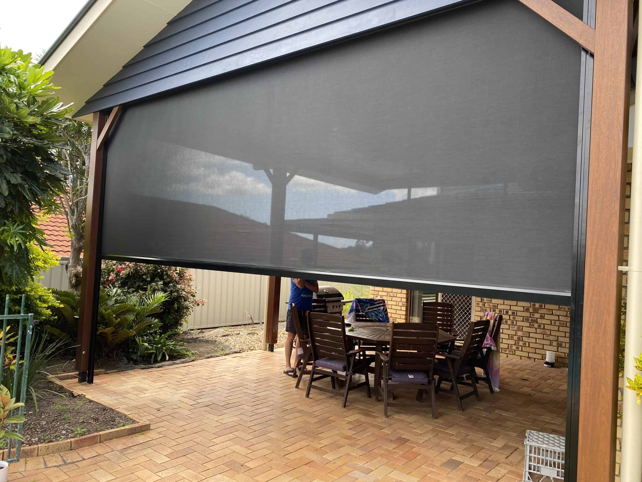 A covered outdoor patio area in Shailer Park features a wooden dining table and chairs, partially enclosed by a large black mesh screen. A person stands nearby, arranging items on the table.