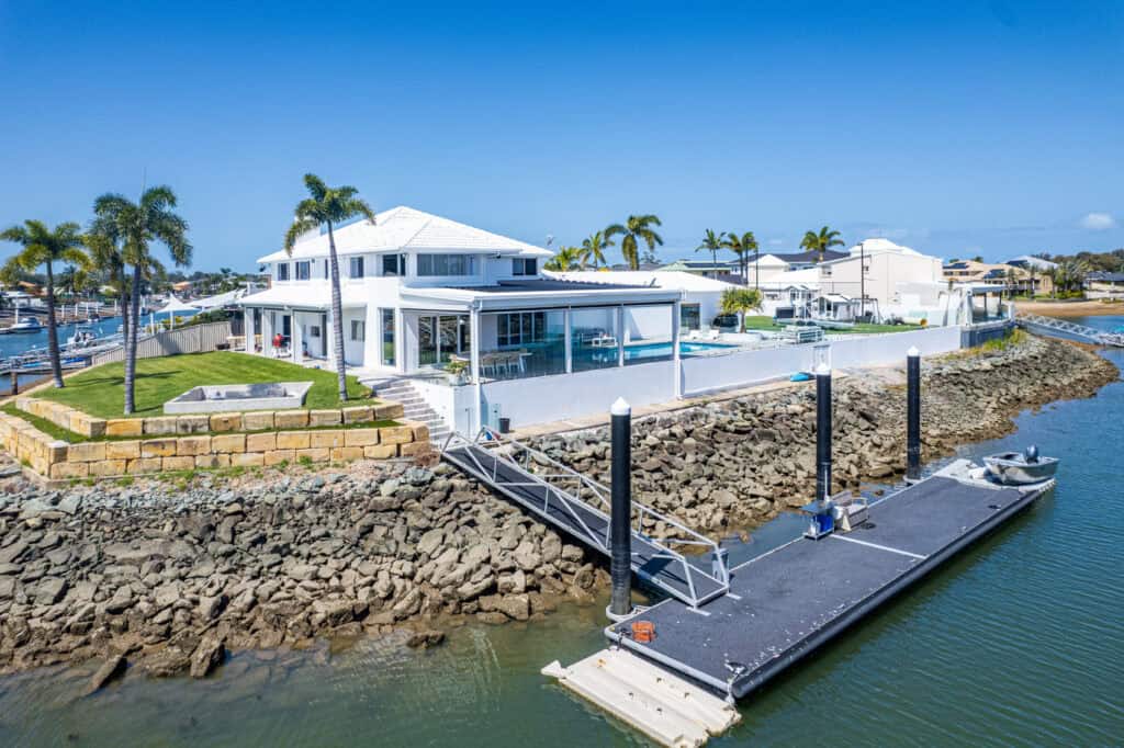 Modern Newport waterfront house with a white exterior, pool, and palm trees, featuring a private dock on a rocky shoreline under a clear blue sky.