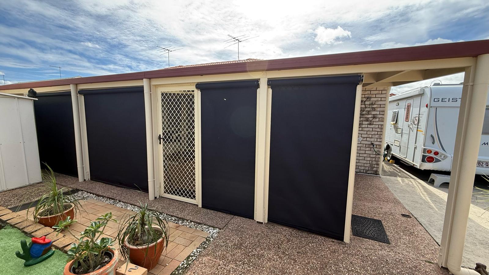 A single-story building with three large dark outdoor blinds, a mesh door, potted plants in front, and a white caravan parked beside it under a partly cloudy sky.