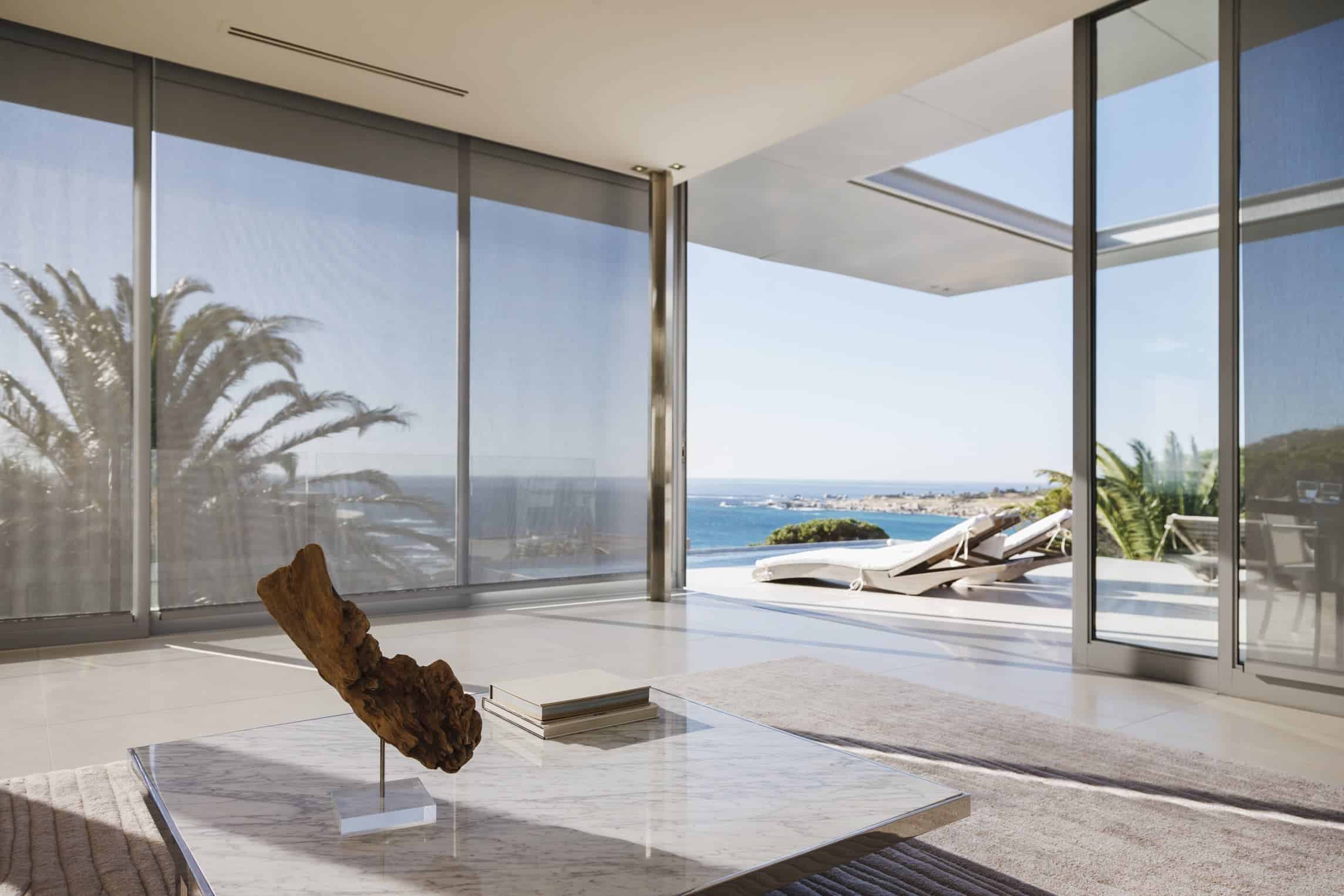 Modern living room with large glass doors open to a patio, showing lounge chairs and a view of the sea and palm trees. A marble coffee table with decor is in the foreground.