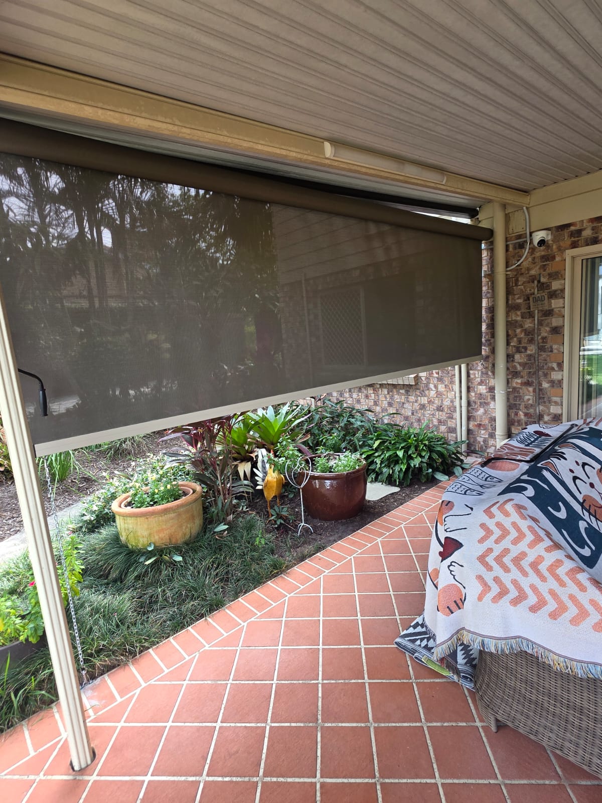 Covered patio with brick flooring, outdoor blinds partially lowered, potted plants, and a couch with a patterned blanket. Garden and greenery visible outside the patio area.