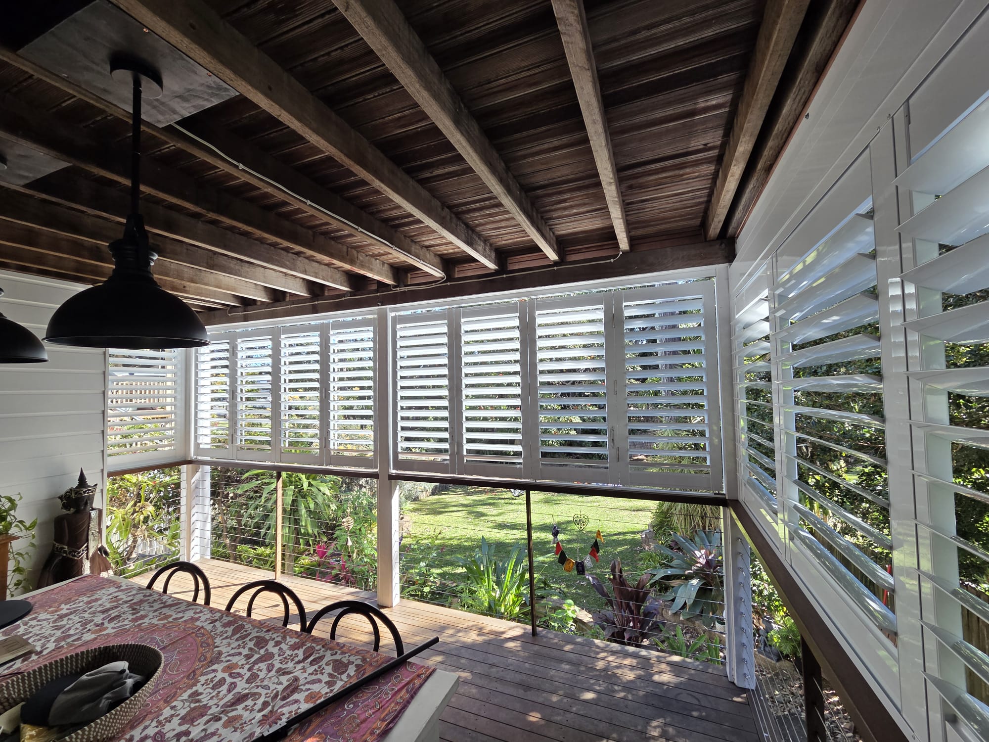 A covered patio with wooden ceiling, white louvered shutters, a table with a patterned cloth, black chairs, and a view of a green garden outside.
