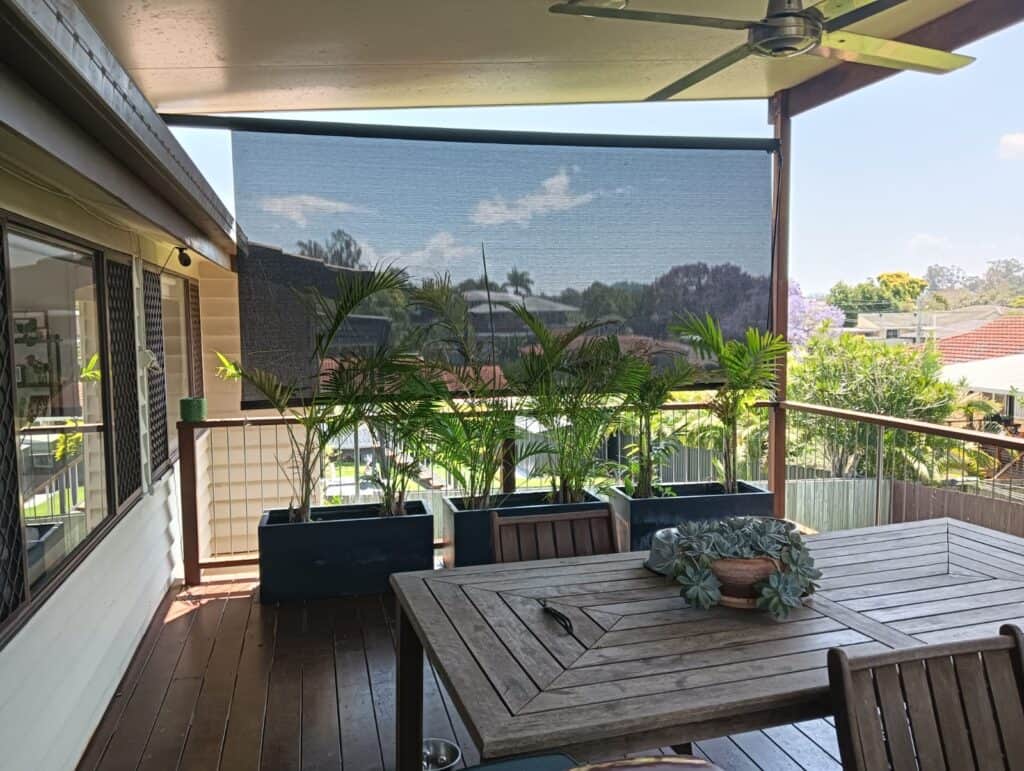 A timber outdoor patio with a table, chairs, potted plants, a ceiling fan and a mesh sunshade overlooking a suburban outlook.