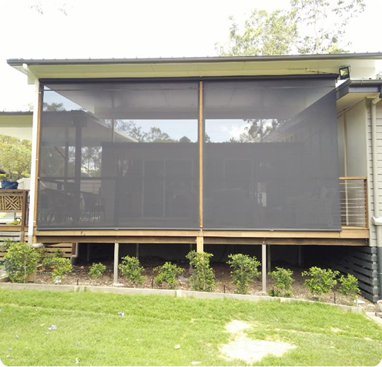 A house patio with two large, dark outdoor roller blinds covers the open area. Small green shrubs line the garden bed below the deck.