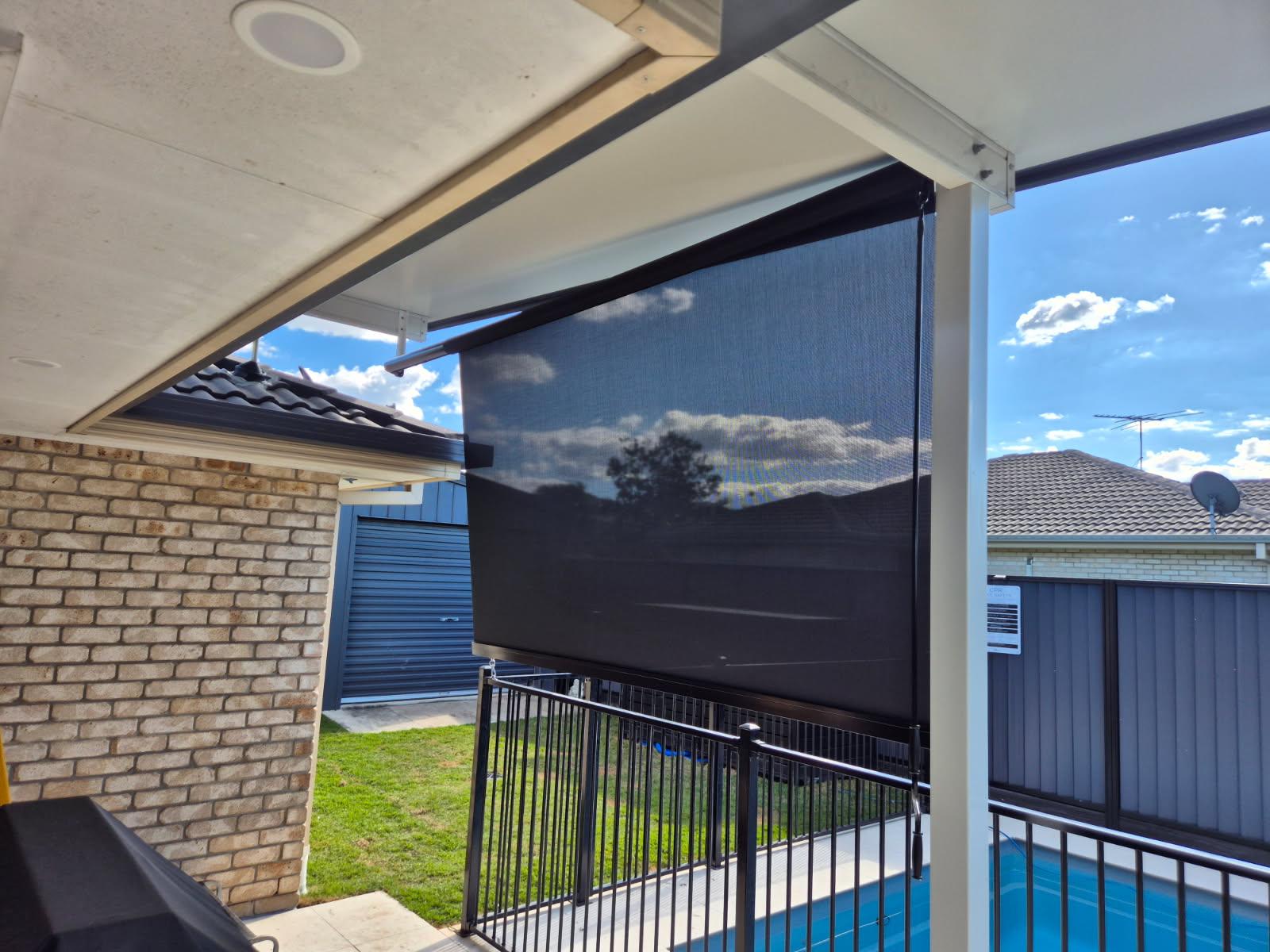 A black outdoor roller shade is partially lowered on a covered patio next to a pool, with neighboring houses and a cloudy sky visible in the background.