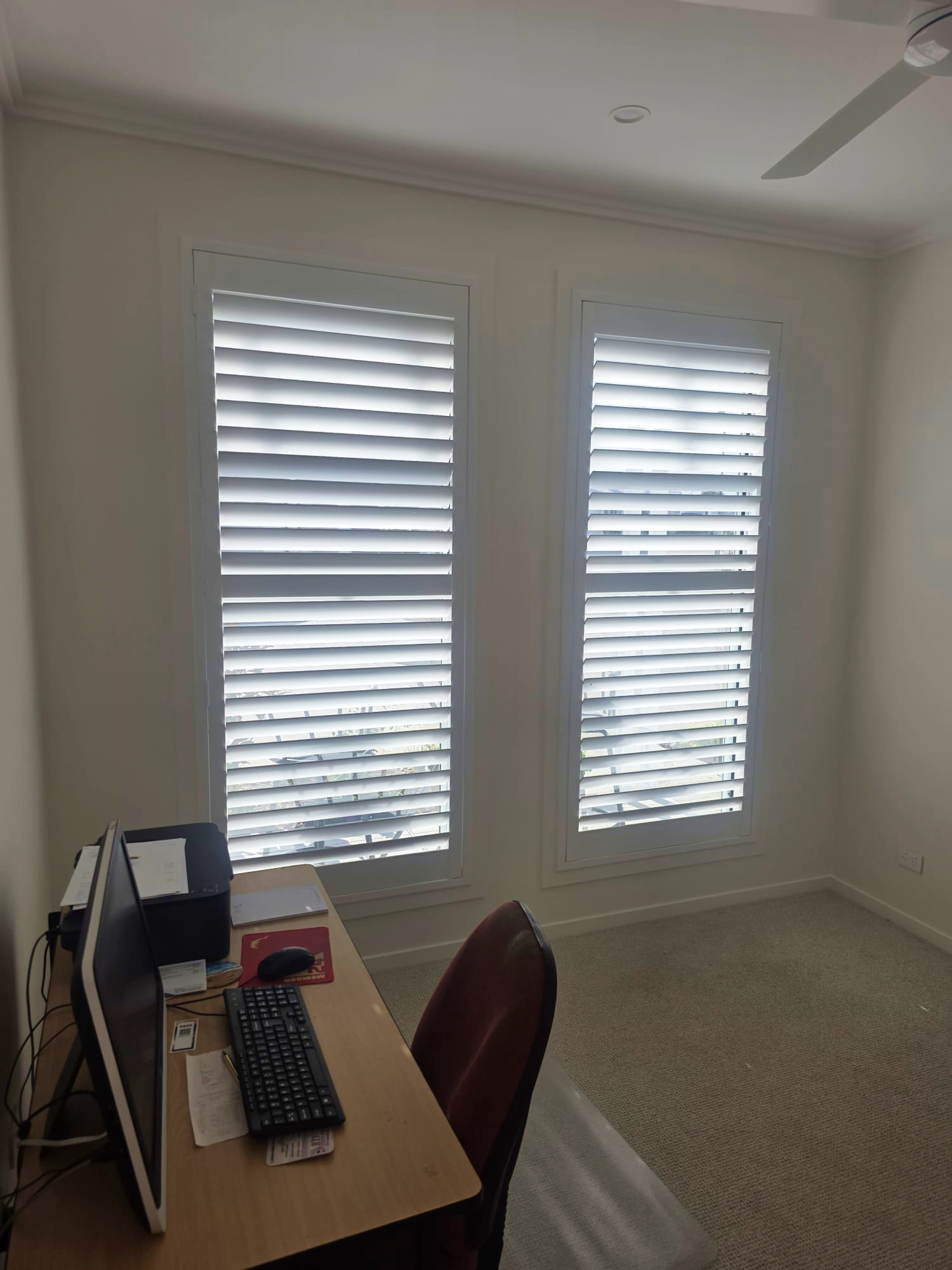 A small office with a desk, computer, printer, and chair is next to two tall windows with white blinds partially closed.
