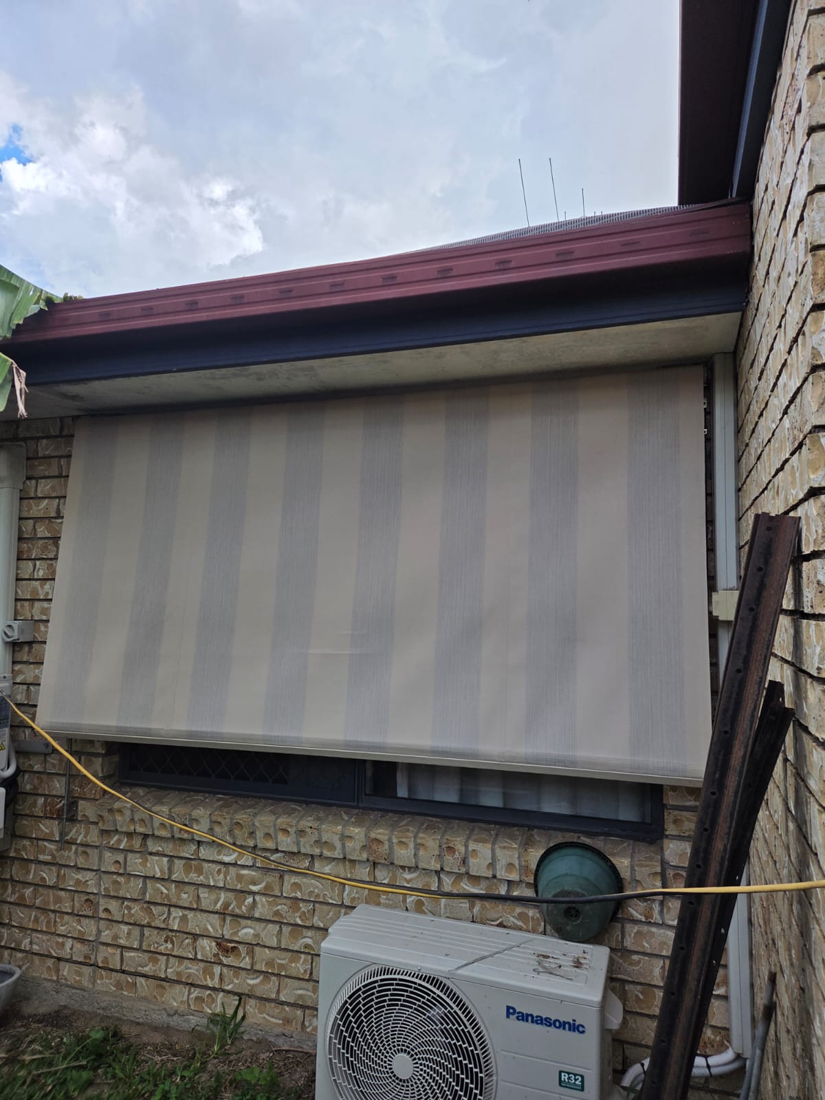 A beige outdoor roller blind covers a window on a brick wall. An air conditioning unit and a hose reel are positioned below the window.