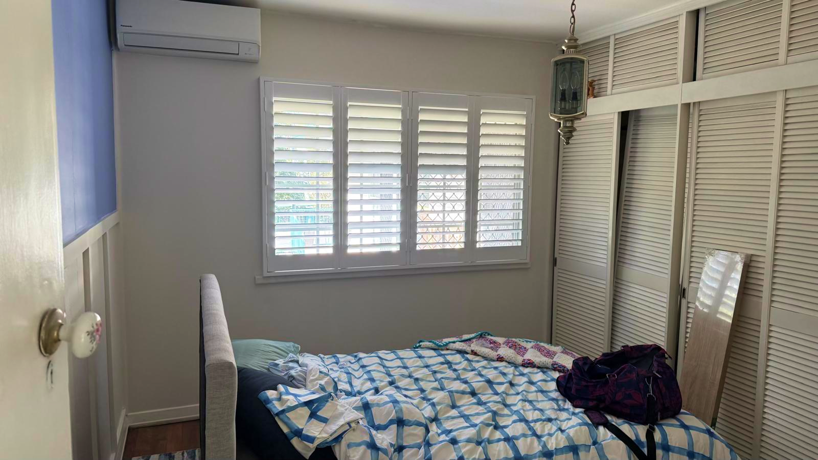 A small bedroom with a bed, rumpled bedding, a bag on the bed, closed shutters on the window, louvered closets, an air conditioner, and a hanging light fixture.