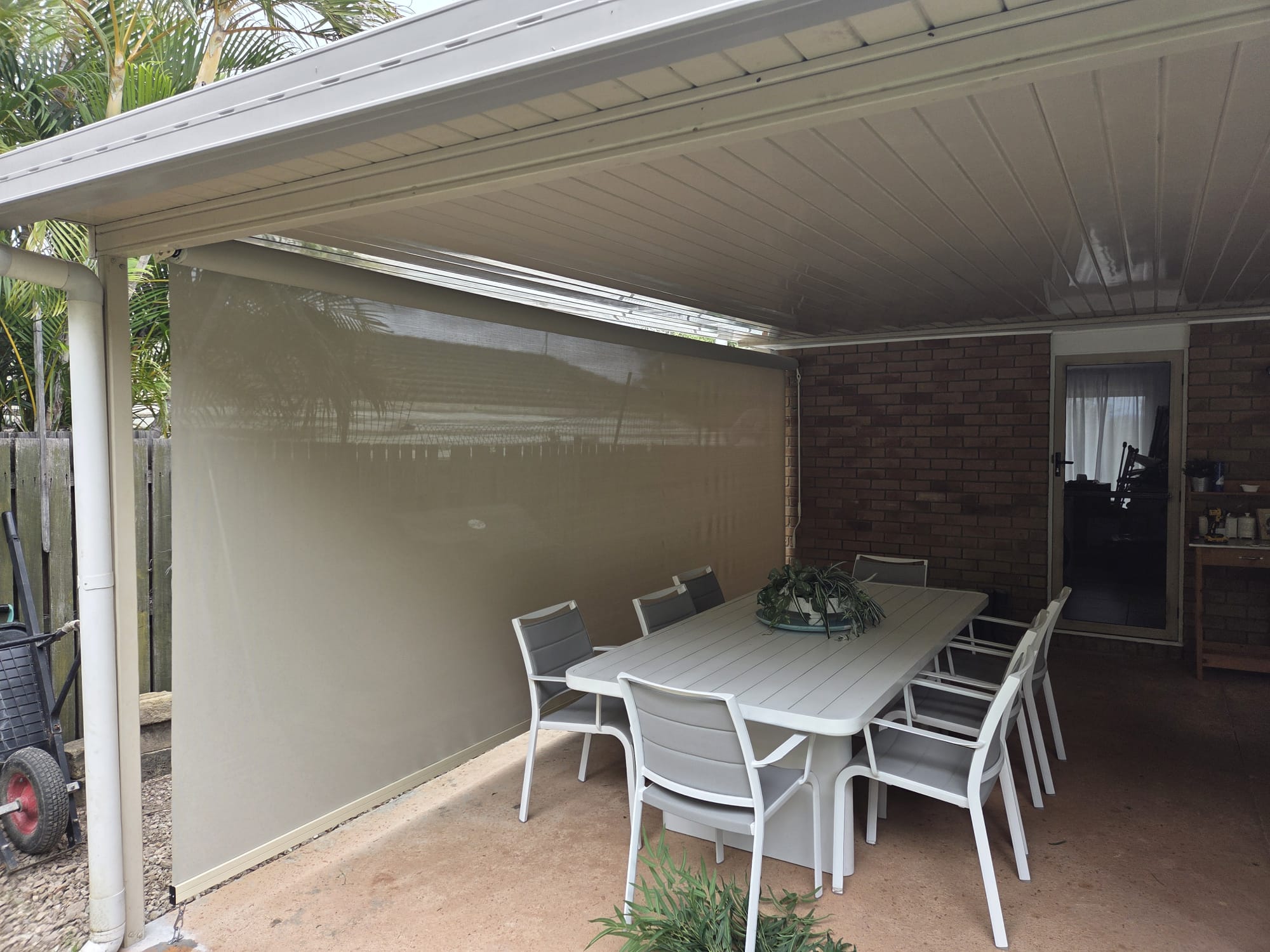 Covered patio area with a long white table, eight chairs, a decorative centerpiece, a beige roll-down shade, and a brick wall with a glass door in the background.