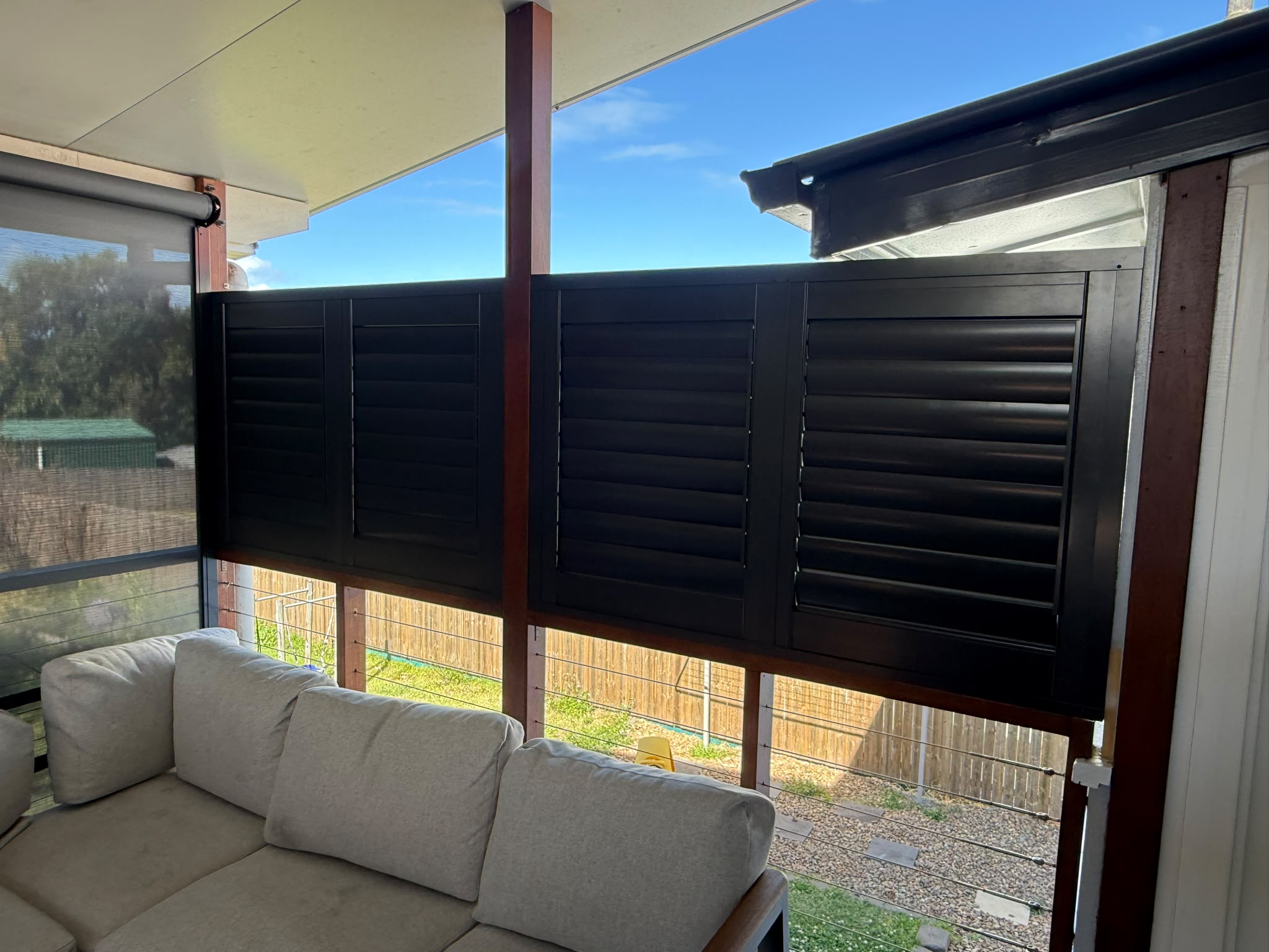 Black louvered shutters are installed on a covered patio above a beige outdoor sofa, overlooking a fenced backyard with grass and garden stones.