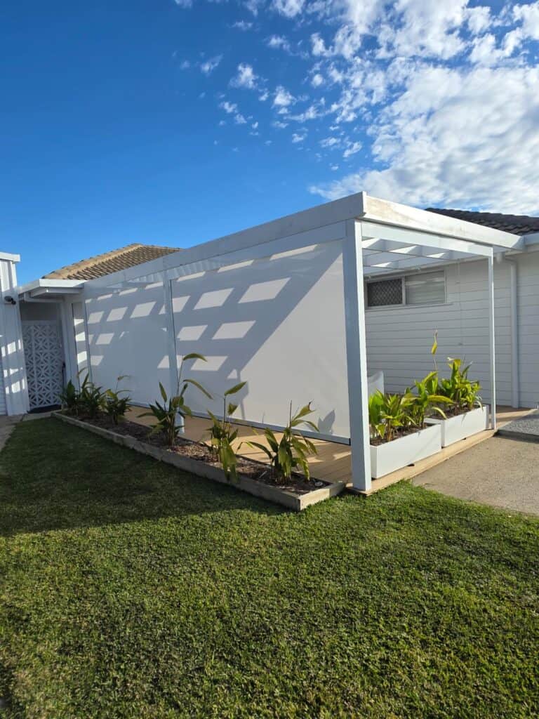 A white pergola with planter boxes and green plants beside a white building, set on a lawn under a partly cloudy sky.