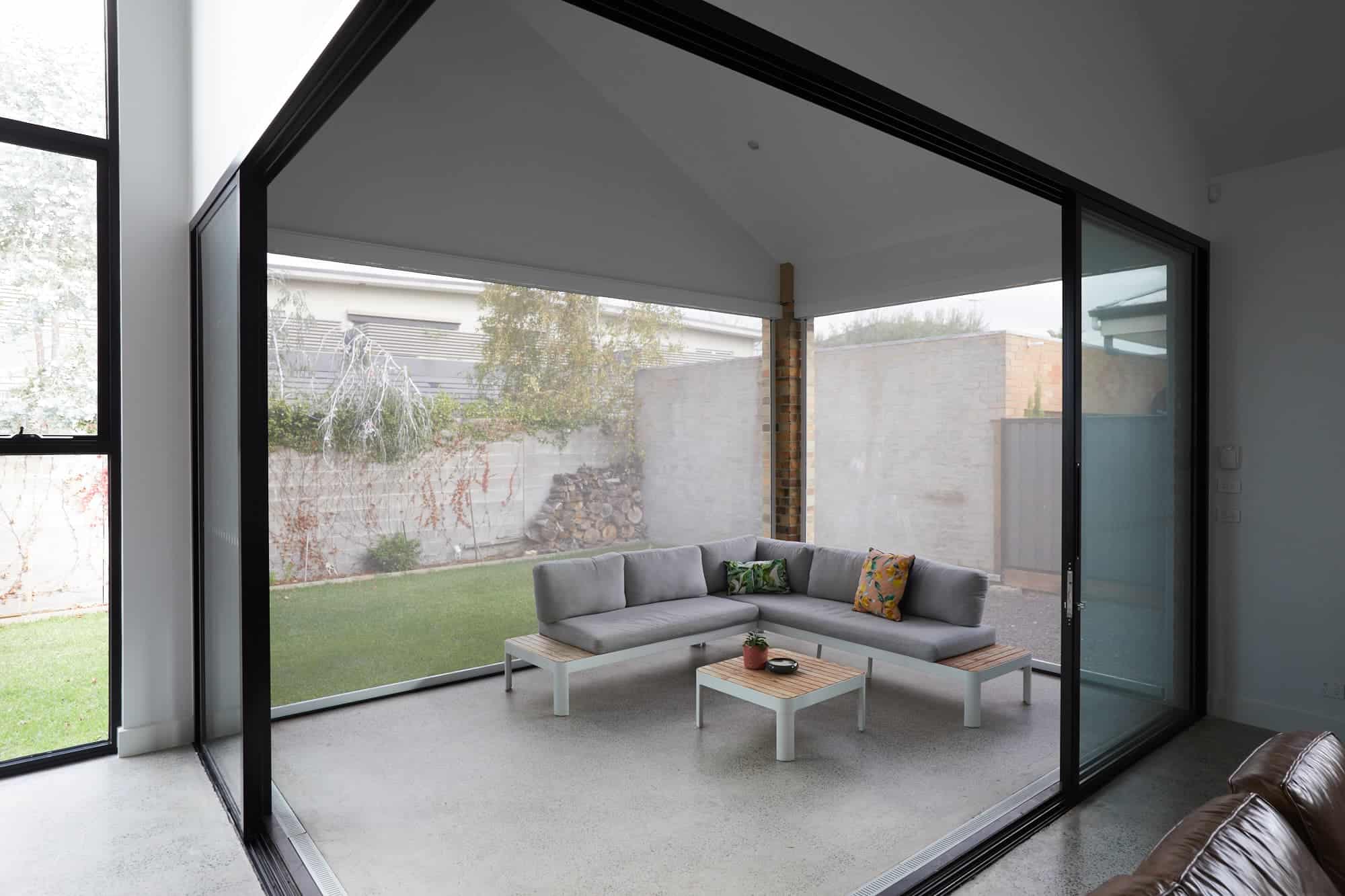A modern glass-enclosed patio features a gray sectional sofa, two small tables, and views of a grassy yard with trees and a brick wall.