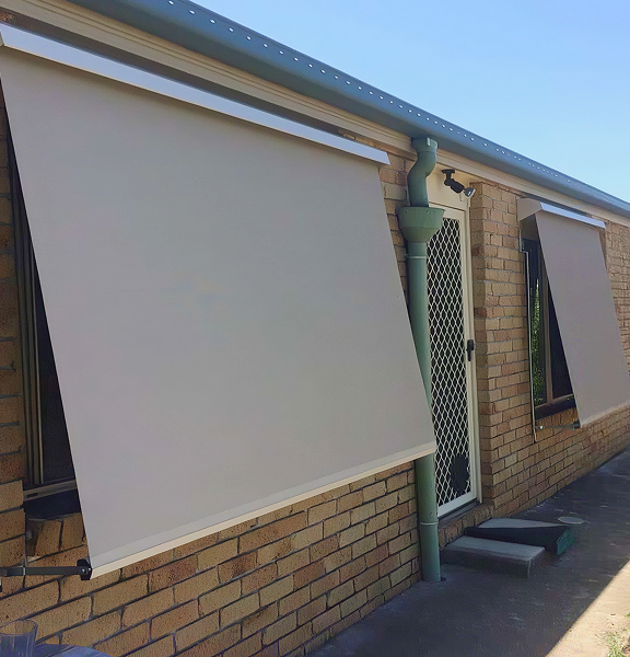 admin-ajax (2) 1 (3) Two beige outdoor roller blinds are extended over windows on a brick house, next to a screen door and a green drainpipe, on a sunny day.