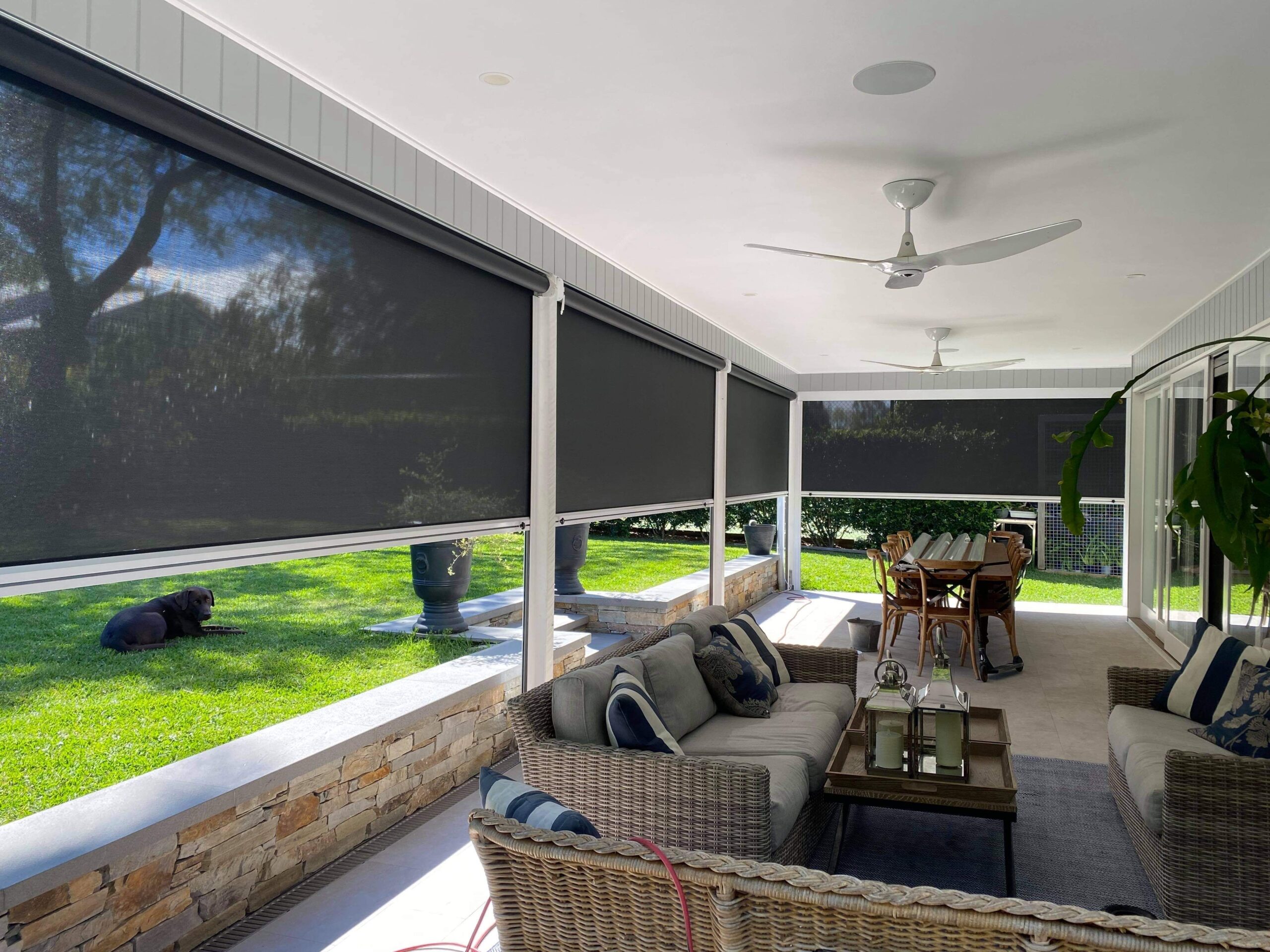 Covered patio with wicker furniture, ceiling fans, and black outdoor roller blinds. A dining table is visible, and a dog lies on the grass outside. Lush greenery surrounds the porch area.