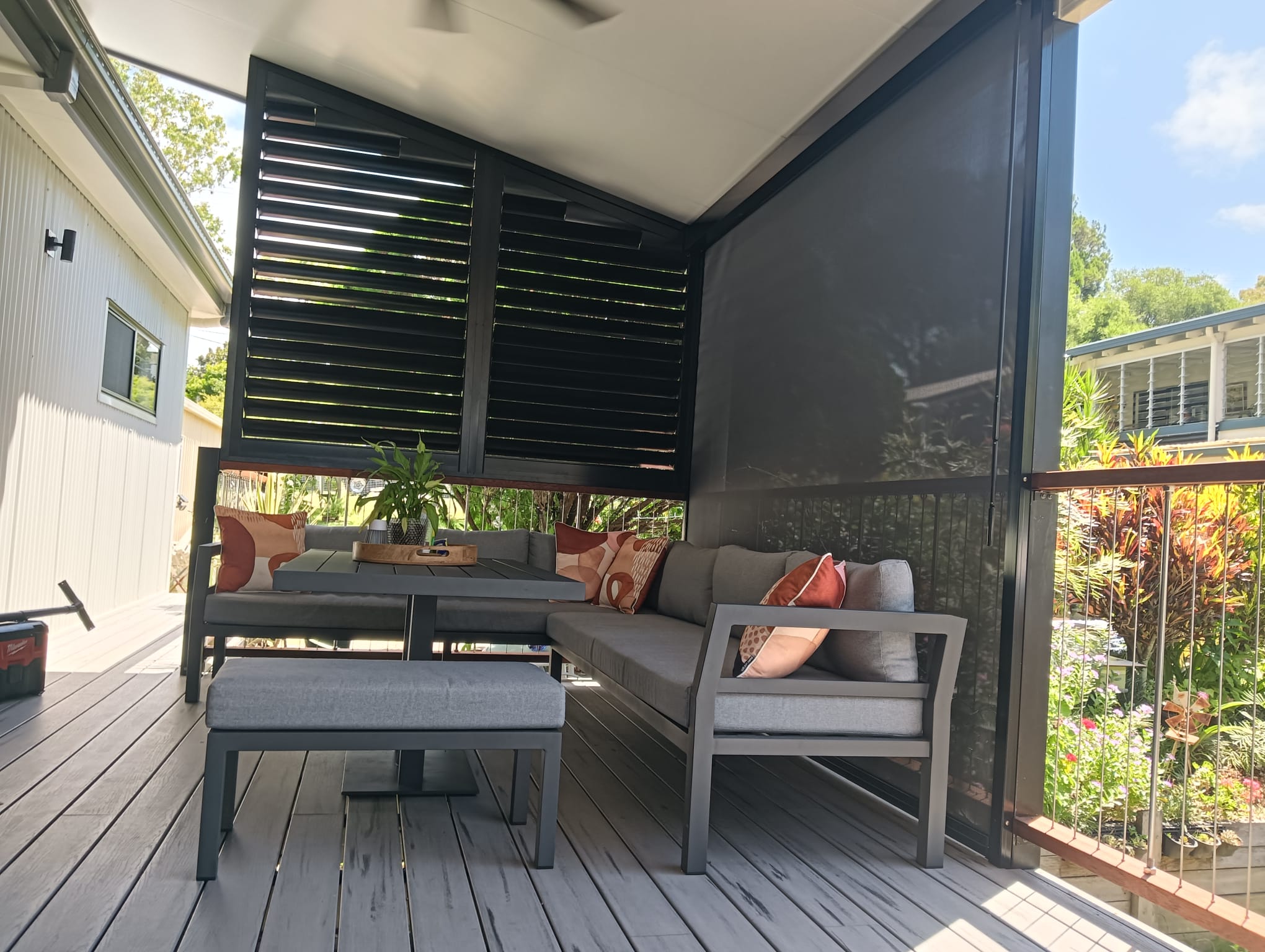 Modern outdoor patio with gray seating, small table, cushions, potted plant, and privacy screens, overlooking a garden with plants and bushes.