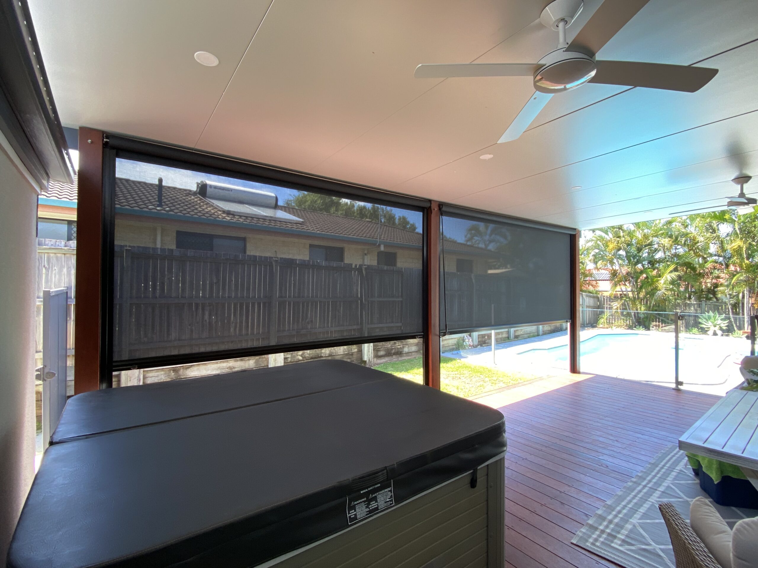 Enclosed patio with ceiling fans, hot tub, outdoor blinds, wooden floor, and view of a backyard with a pool, fence, and house.