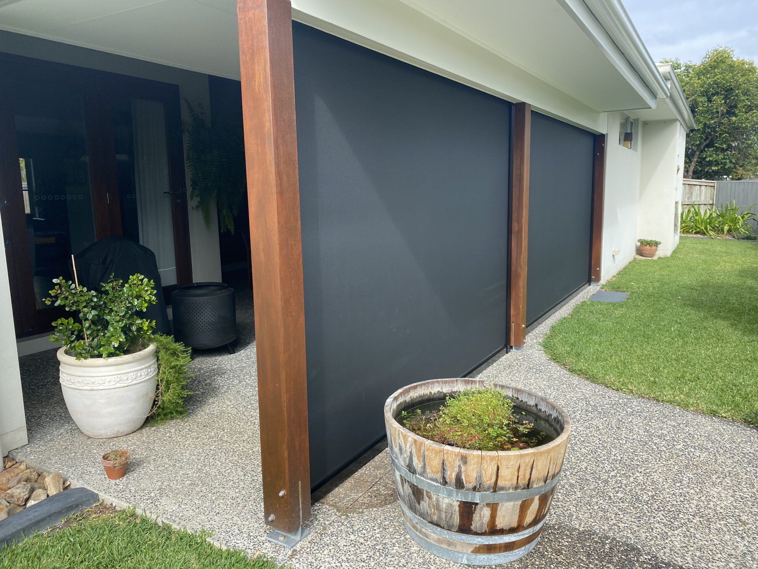 Outdoor patio with wooden posts, black roller blinds, potted plants, and a round water barrel on a pebble path next to a grassy lawn.