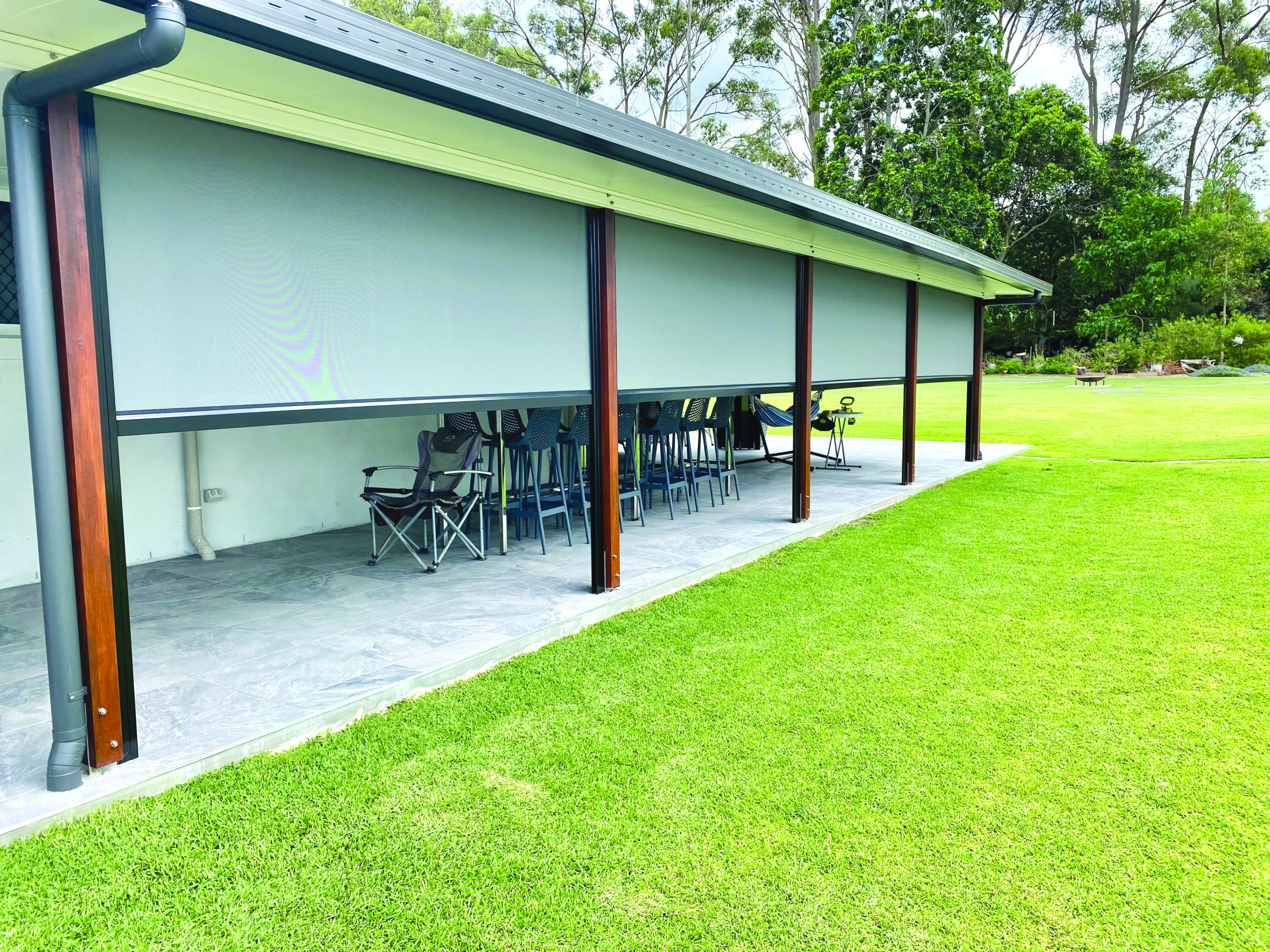 Outdoor covered patio area with chairs and tables, partially enclosed by a large pull-down screen, adjacent to a grassy lawn and surrounded by trees.