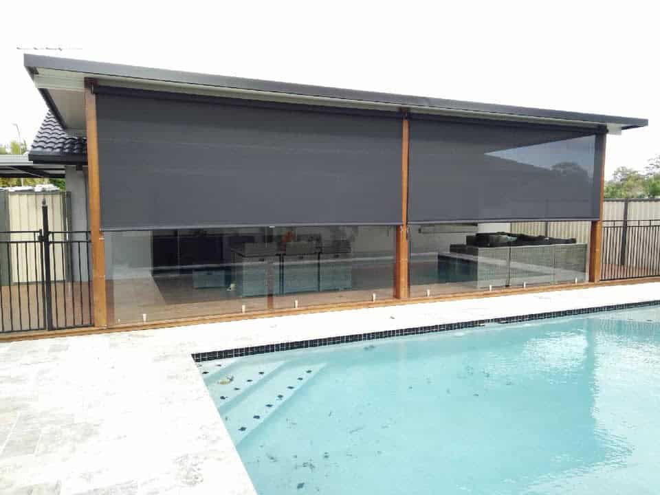Outdoor pool area with a glass fence in Burpengary, adjacent to a covered patio featuring lowered dark roller shades.