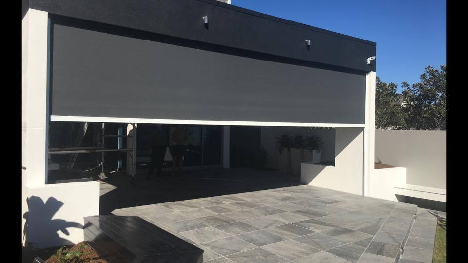 A modern Albany Creek house with a large grey retractable patio screen partially lowered over a tiled outdoor area, under a clear blue sky.