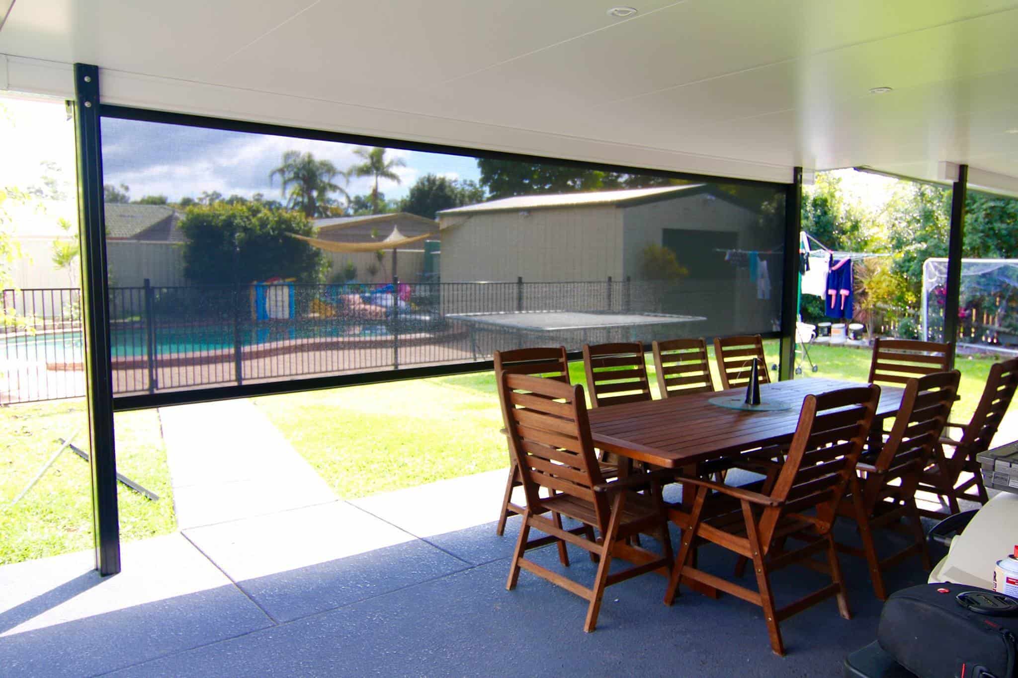 Covered patio area with a long wooden dining table and chairs, overlooking a backyard with a pool, garden, and laundry hanging outside. A shade screen is partially lowered—perfect for relaxed Manly living.