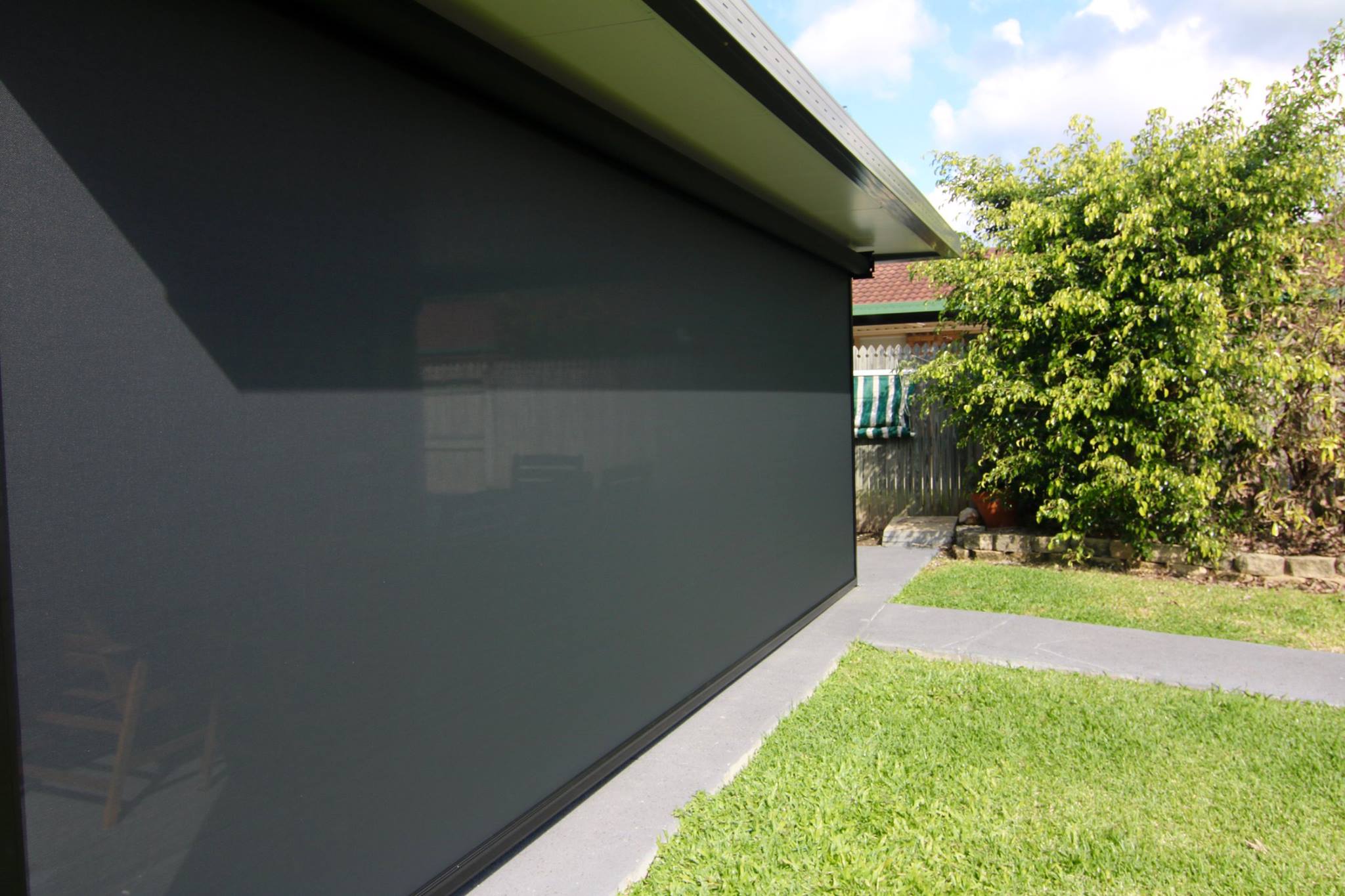 Outdoor roller shade fully drawn down along a patio area, with a grassy lawn, tree, and a fence in the background under a partly cloudy sky.