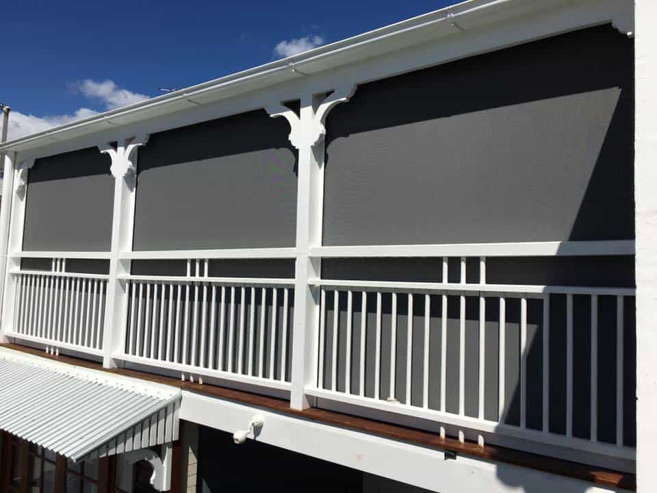 A white balcony in North Lakes with vertical railings and decorative supports features large grey outdoor roller blinds covering the upper section.