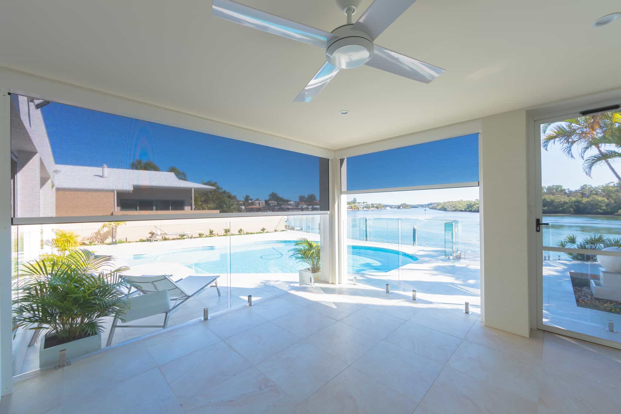 A spacious covered patio with a ceiling fan overlooking a pool, featuring glass railings, loungers and partially lowered blue roller blinds.